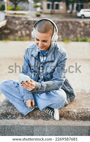 Young bald woman listening music playlist while using mobile phone outdoor - Main focus on eyes - Vertical photo