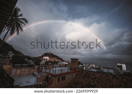 From a favela, a wide-angle low-key view of a balcony facing a backdrop of dark, moody skies; a beautiful rainbow shines through the clouds, adding a burst of color and hope to the scene;