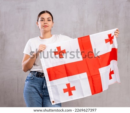 Confident young woman in casual wear holding flag of Georgia against gray background indoors