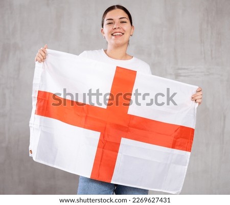Joyous young woman with England flag in hands posing happily against light unicoloured background