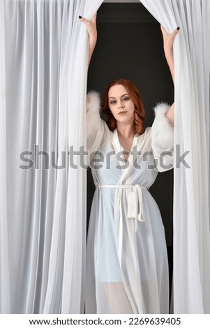 Portrait of beautiful red haired woman wearing glamorous  silk  bridal dressing gown. Standing pose in front of white curtains in bedroom background. 