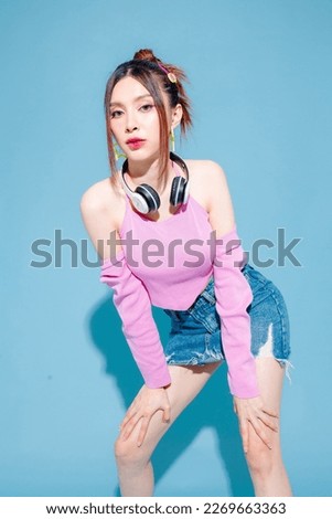 Young fashionable cute carefree Asian woman wearing headphone and roller skate with perfect slim body on isolated blue background. Positive model having fun and cool posing indoors studio.