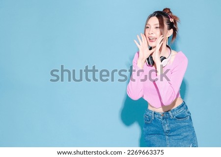 Young fashionable cute carefree Asian woman wearing headphone and roller skate with perfect slim body announce on isolated blue background. Female model posing indoors studio.