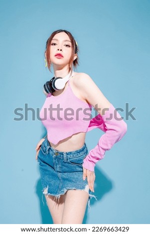 Young fashionable cute carefree Asian woman wearing headphone and roller skate with perfect slim body on isolated blue background. Positive model having fun and cool posing indoors studio.