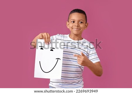 Little African-American boy holding paper with happy emoticon on pink background