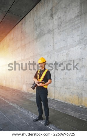 Asian engineer wearing hard hat and safety vest looks at the camera with a serious look while holding tablet and hang up camera in construction site.