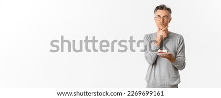 Image of thoughtful and serious middle-aged man, celebrating birthday, holding b-day cake and thinking what wish to make, standing over white background.