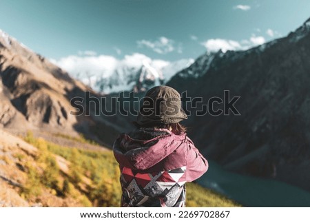 A traveler girl in a hat takes pictures with her back to the camera on the phone of an alpine lake with mountains with snow and glaciers in Altai.