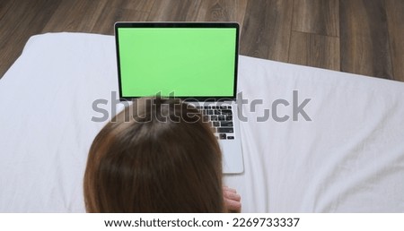 Woman works behind laptop in lying on a bed on a white sheet, top view. Green screen, chroma key, typing on the keyboard, touchpad. Concept of remote work, study, comfortable workplace.