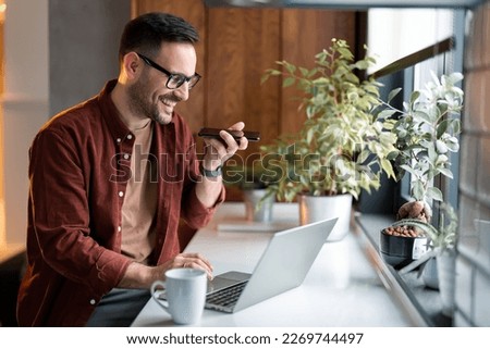 Satisfied modern man in stylish casual clothes enjoying a pleasant conversation on smart cell phone while spending time at home. Happy millennial man talking on mobile phone and using laptop computer. Royalty-Free Stock Photo #2269744497