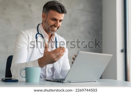 Happy encouraging male doctor physician talking, consulting patient online by webcam video call on laptop computer. Confident doctor on telemedicine conference virtual tele meeting in hospital office.