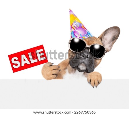 French bulldog puppy wearing sunglasses and party cap shows signboard with labeled "sale" above empty white banner. isolated on white background