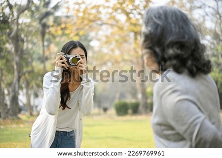Beautiful and happy Asian granddaughter taking a picture of her grandmother with retro camera while spending time together in the beautiful green park.