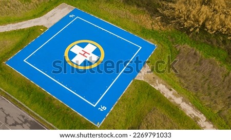 Aerial closeup of a helicopter landing pad near emergency hospital and medical centre. A blue asphalt-covered helipad with a special symbol in the center for helicopter landing, near a green field. 