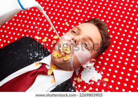 Young man lying on red vintage table cloth with corn flakes in mouth. Milk pouring inside mouth. Breakfast time. Food pop art photography. Complementary colors. Copy space for ad, text