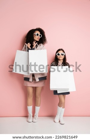 Full length of trendy woman in sunglasses holding shopping bags near child on pink background