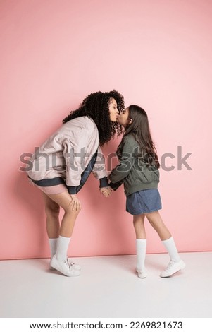 Side view of curly woman kissing stylish daughter on pink background