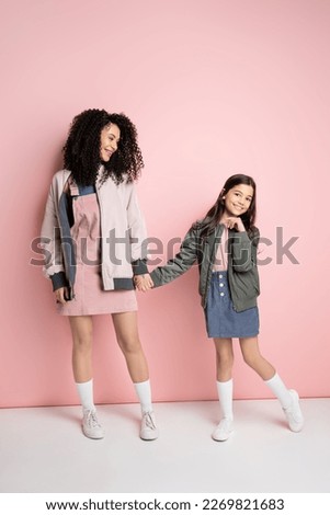 Fashionable girl holding hand of curly mom on pink background