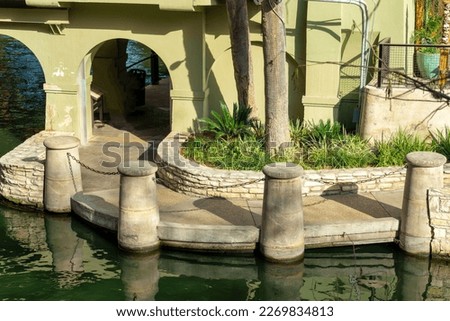 Water canal with cement pillars and chains with archways trees in late afternoon shade and grass palm trees in city. In urban part of san antonio texas cassic and historic waterways for boat tours. Royalty-Free Stock Photo #2269834813