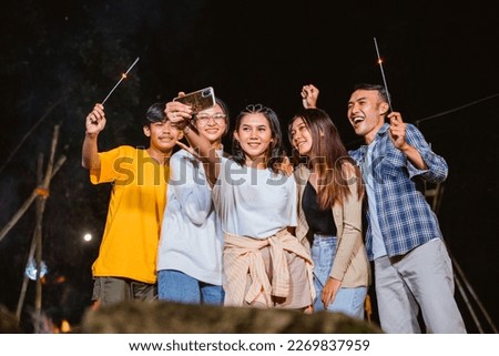 woman in white t shirt standing at the center and holding the phone while taking groufie photo with her friends at the camp site