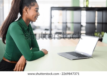 Young african-american female student coworker sitting at the desk in modern coworking space in front of laptop, looking at mock up white computer screen. Over shoulder back view