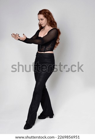 Full length portrait of beautiful woman with long red hair wearing black corset top and leather pants. Standing pose, walking forwards with gestural hands reaching out. Isolated on white studio backgr