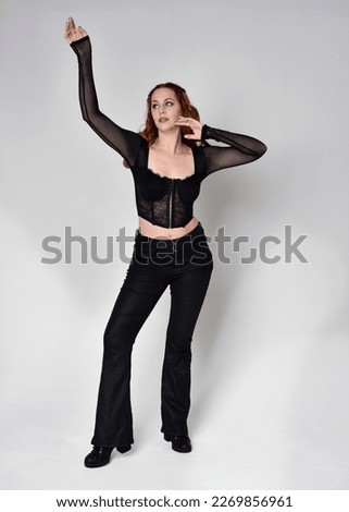 Full length portrait of beautiful woman with long red hair wearing black corset top and leather pants. Standing pose, walking forwards with gestural hands reaching out. Isolated on white studio backgr