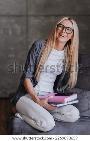 Cheerful blonde in glasses fashion model relaxing home, sitting with book on cozy chair happily against modern interior. Successful businesswoman relaxing after hard week. Purposeful female.