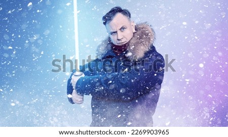 Man in a winter jacket without a hat with a light sword in his hands on a blue background with falling snow.
