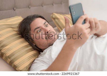 A chubby man in his 30s is shocked by a controversial social media post on his cellphone while lying on the bed. Royalty-Free Stock Photo #2269921367