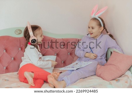 two sister girls have fun at home, draw a bunny and listen to music in headphones, the older girl has a hairband with rabbit ears on her head, waiting for the Easter holiday