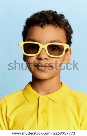 On style. Closeup portrait of little african cute boy in stylish sunglasses and yellow summer shirt looking at camera over blue background. Concept of facial expression, happiness, kids emotions
