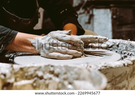 hands young woman close-up are stained in clay, they make ceramic product on potter's wheel. Close-up, soft focus, against blurred background of pottery workshop. Potter's hands at work