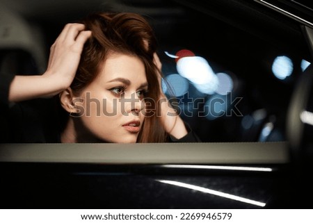 horizontal photo from the side, at night, of a woman sitting in a car and looking out of the window, straightening her hair with her hands