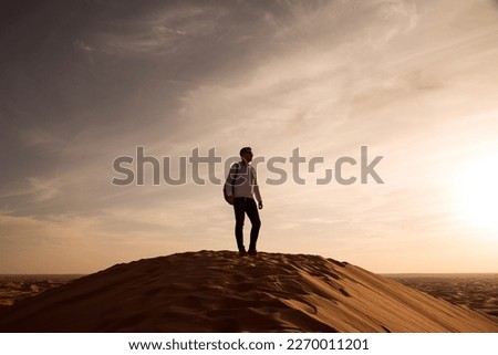 
silhouette of a man with a backpack standing on a dune at sunset. Royalty-Free Stock Photo #2270011201