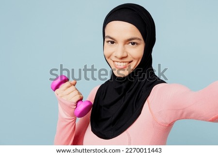 Young asian muslim fitness trainer sporty woman wear pink abaya hijab spend time in gym do selfie shot mobile cell phone hold dumbbell isolated on plain blue background. Workout sport fit abs concept