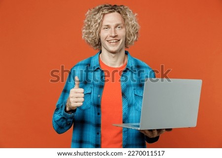 Young smiling smart blond caucasian IT man wears blue shirt orange t-shirt hold use work on laptop pc computer show thumb up isolated on plain red background studio portrait. People lifestyle concept