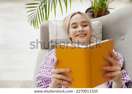 Young smiling happy smart woman wearing casual clothes reading book novel lying on grey sofa couch stay at home hotel flat rest relax spend free spare time in living room indoor. People lounge concept