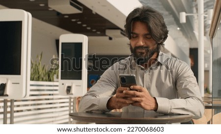 Happy young indian businessman holding smartphone sending message sitting indoors smiling arabian man play mobile game using application on phone reading good news on internet chatting in social media