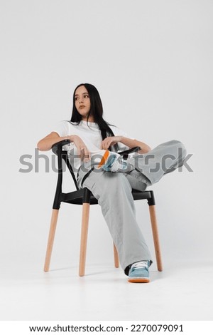 Portrait of a beautiful brunette girl sitting on a chair, dressed in a white shirt, wide jeans and sneakers. Isolated on white. Royalty-Free Stock Photo #2270079091