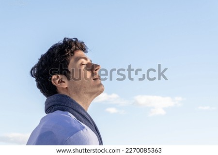 Young curly haired boy isolated on background of clear sky and with closed eyes smelling blue sky. Royalty-Free Stock Photo #2270085363