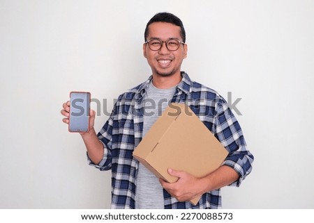 Adult Asian man smiling and showing blank mobile phone screen while holding a package cardboard