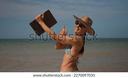 Pretty happy smiling mature senior woman in hat sunglasses and bikini holding laptop at the beach giving thumbs up as digital nomad in tropical place.