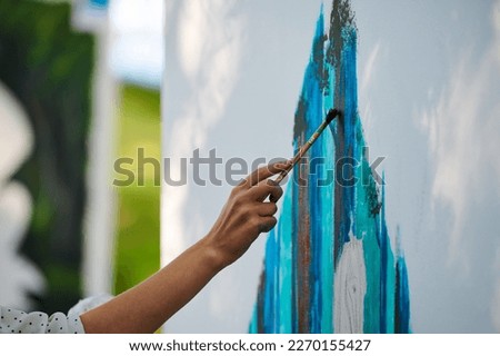 Artist hand holds paint brush and draws abstract surreal image on white canvas at outdoor art painting festival, paintings art picture process. Artist paints atmospheric picture