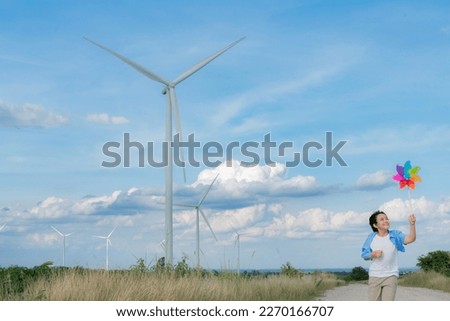 Progressive young asian boy playing with wind pinwheel toy in the wind turbine farm, green field over the hill. Green energy from renewable electric wind generator. Windmill in the countryside concept Royalty-Free Stock Photo #2270166707