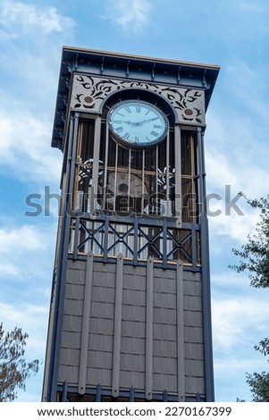 Modern decorative clock tower with square shade and metal engineered facade with white cloudy blue sky background. Late afternoon shade with visible trees in park outdoor city background.