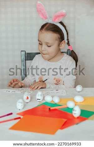 a little girl in bunny ears on her head decorates white eggs with stickers with different emotions, in her hands is a white egg with emotion