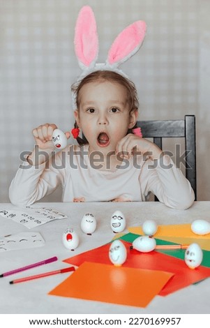 a little girl in bunny ears on her head decorates white eggs with stickers with different emotions, in her hands is a white egg with emotion, the child copies and shows with facial expressions.