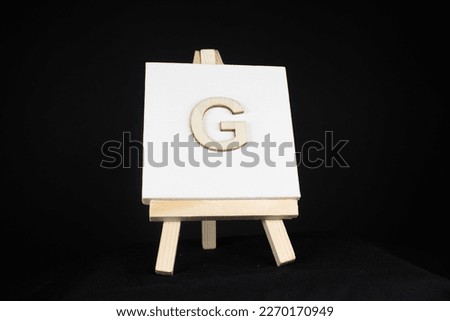 G wooden capital letter and white blank painting canvas resting on an artists easel isolated on a black background