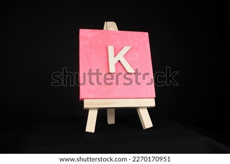K wooden capital letter and pink blank painting canvas resting on a miniature artists easel isolated on a black background